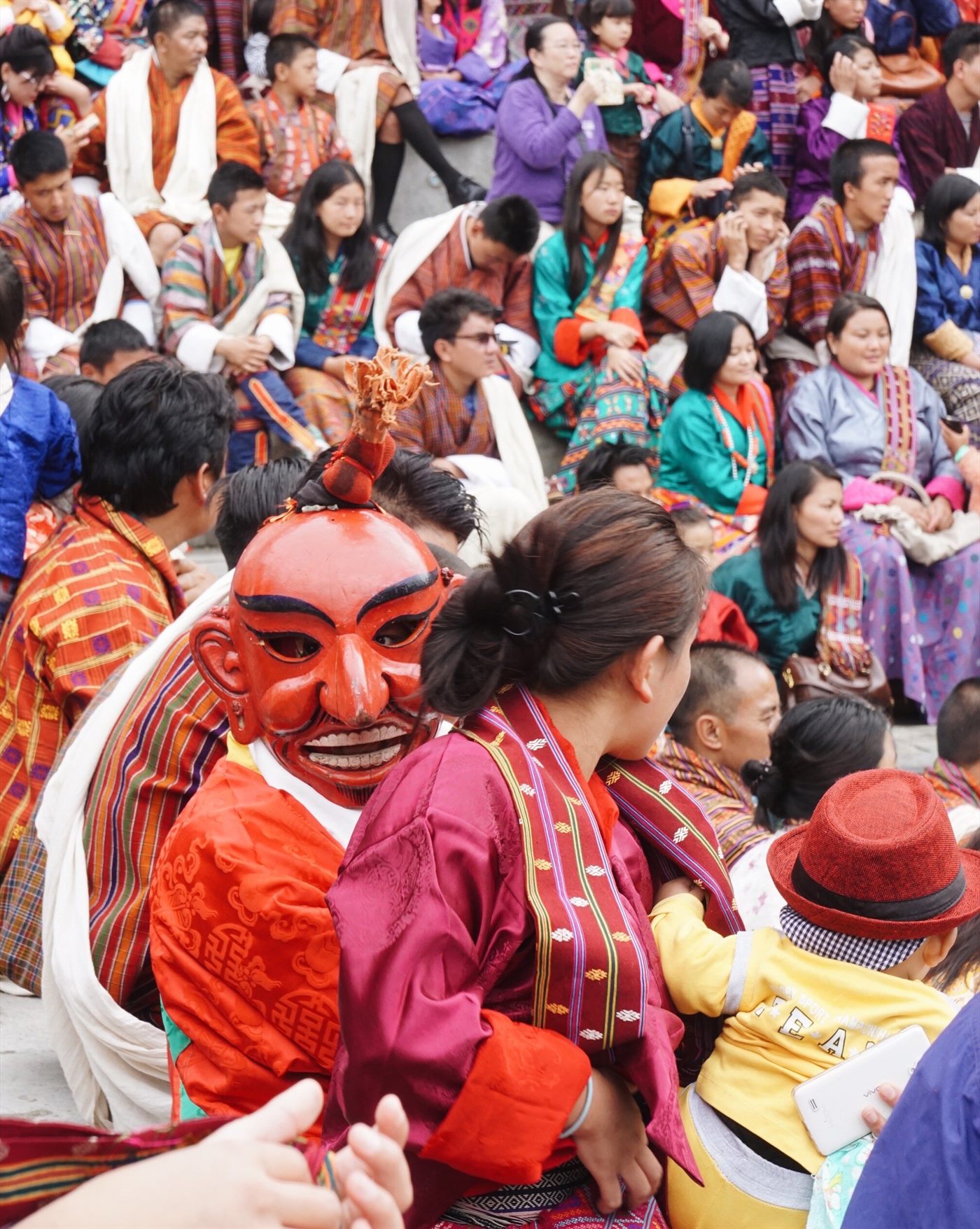 A masked dancer seated among a crowd in traditional dress at a Bhutanese festival