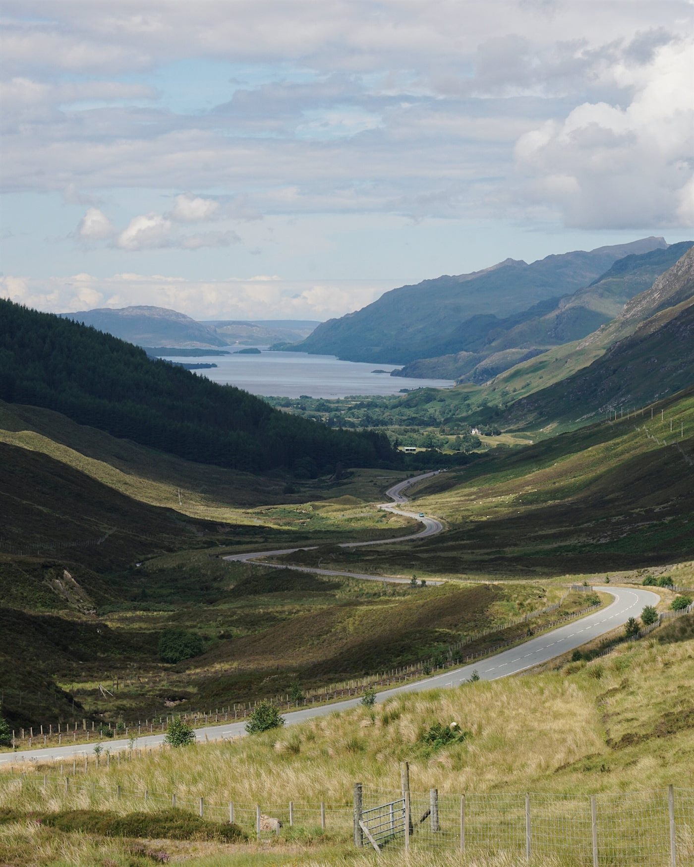 A single-track road winding through a green Highland glen toward a loch, Scotland